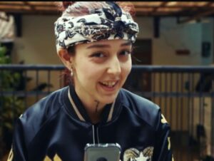 Young woman wearing a patterned headscarf and dark jacket smiles at the camera, holding a phone.