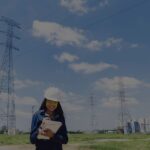 Woman in a white hard hat and yellow safety glasses writing in a notebook at a rural power-transmission site with tall lattice towers and a distant city skyline.