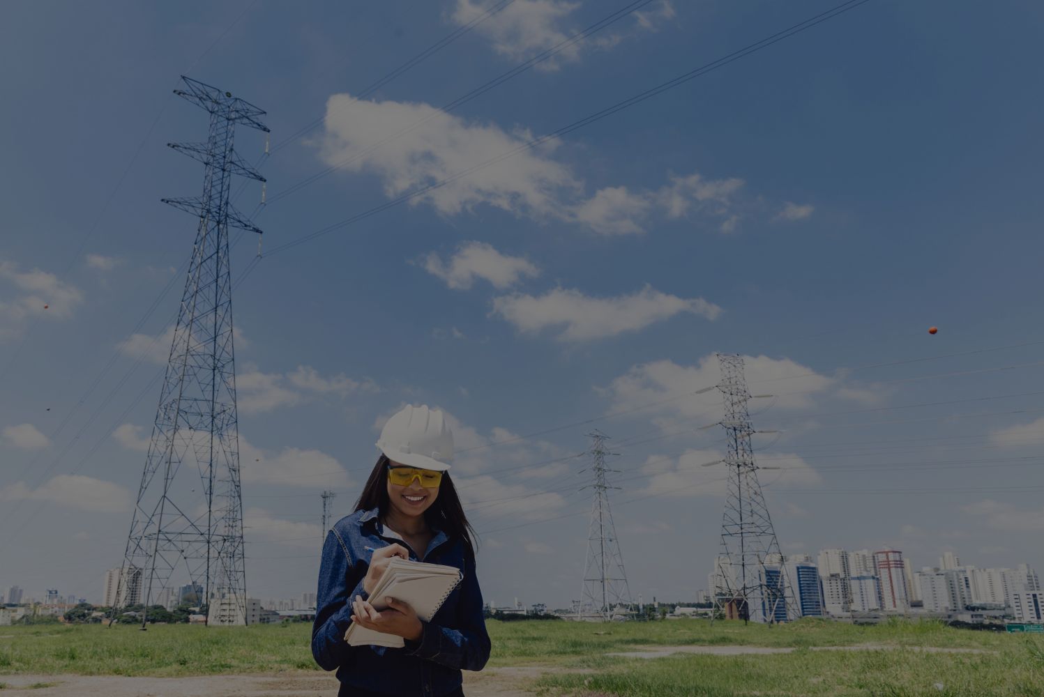 Woman in a white hard hat and yellow safety glasses writing in a notebook at a rural power-transmission site with tall lattice towers and a distant city skyline.