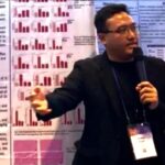 Male presenter in a black blazer speaks into a microphone in front of research posters with red bar charts behind him.