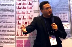 Male presenter in a black blazer speaks into a microphone in front of research posters with red bar charts behind him.