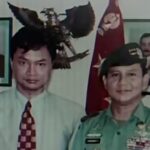 Two men posing for a photo: one in a light shirt and patterned tie, the other in a green military uniform with medals, with portraits and a flag in the background.