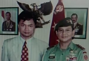 Two men posing for a photo: one in a light shirt and patterned tie, the other in a green military uniform with medals, with portraits and a flag in the background.