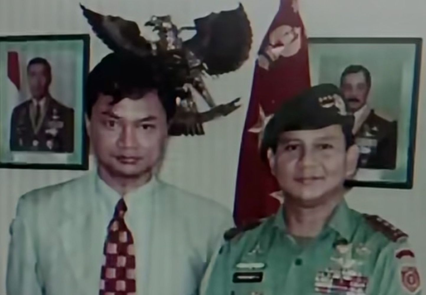 Two men posing for a photo: one in a light shirt and patterned tie, the other in a green military uniform with medals, with portraits and a flag in the background.