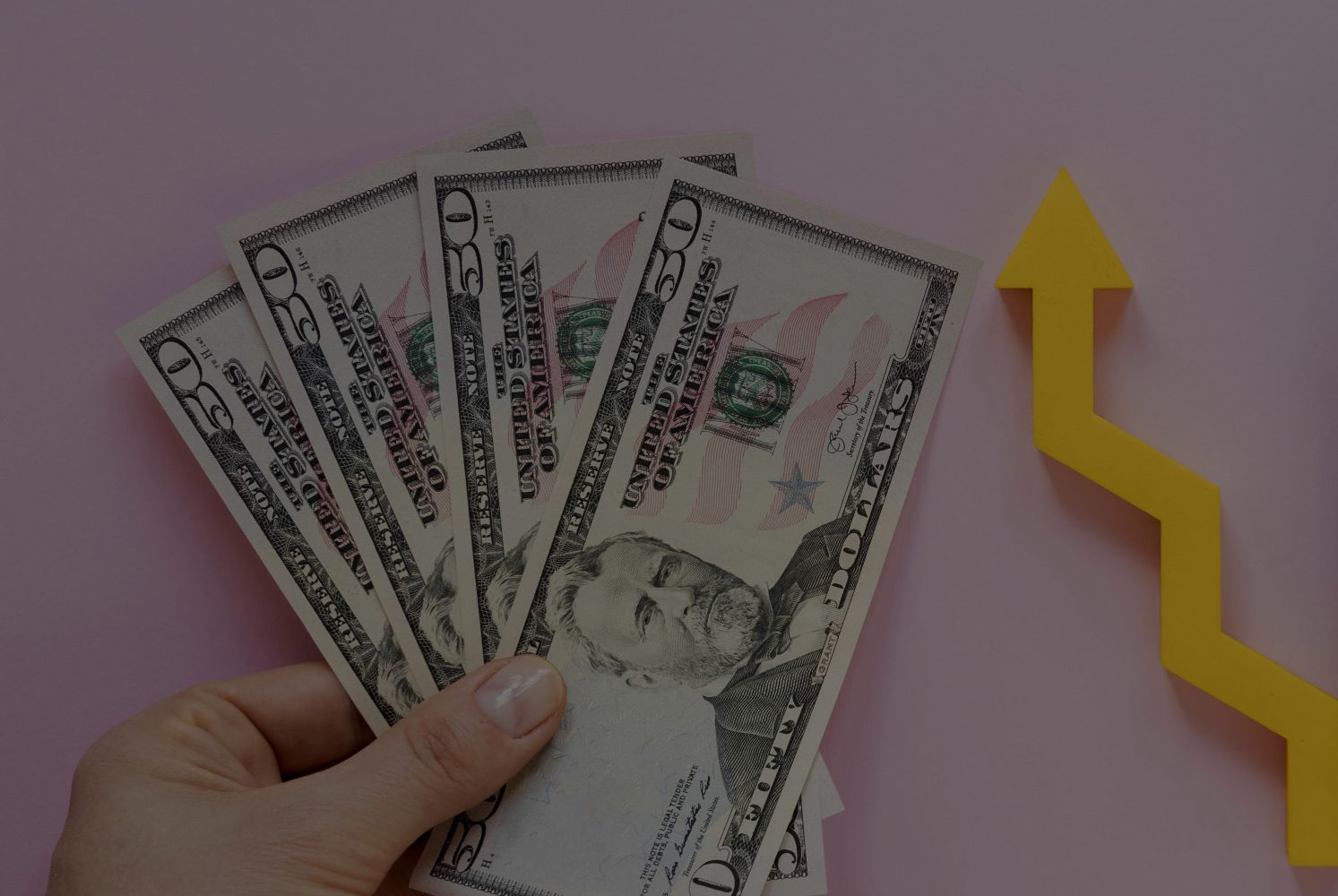 Fanned stack of $50 bills held in a hand with a yellow upward arrow, symbolizing financial growth.