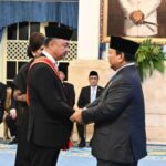Two men in suits wearing traditional black caps shake hands during a formal ceremony, with seated officials in the background and a portrait on the wall.
