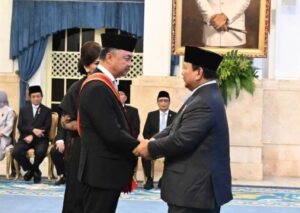 Two men in suits wearing traditional black caps shake hands during a formal ceremony, with seated officials in the background and a portrait on the wall.