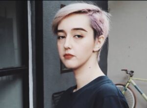 Portrait of a person with short pastel pink and blonde hair, standing near a doorway with a bicycle in the background.