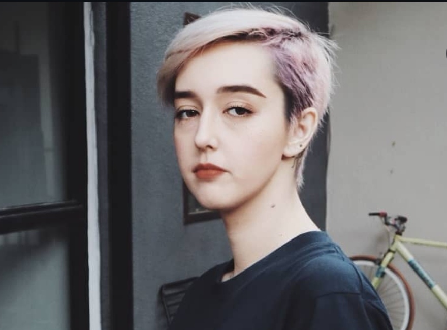 Portrait of a person with short pastel pink and blonde hair, standing near a doorway with a bicycle in the background.