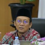 Asian man in a batik shirt and peci speaks at a panel with a microphone, laptop, water bottle, and flowers nearby.