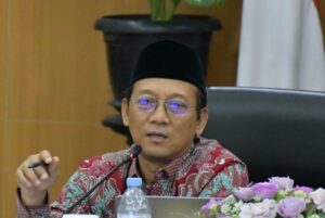 Asian man in a batik shirt and peci speaks at a panel with a microphone, laptop, water bottle, and flowers nearby.