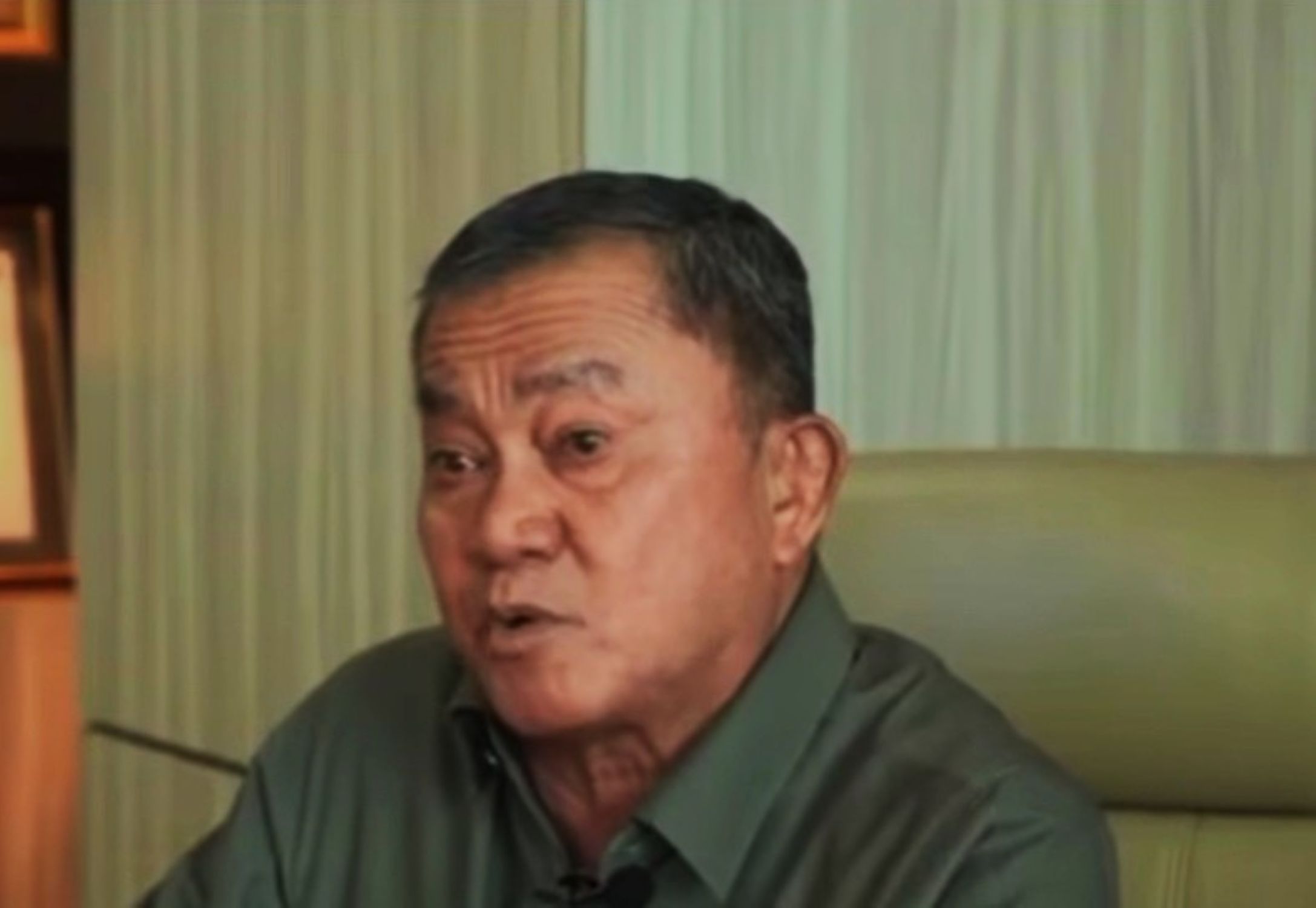 Man in a dark green shirt seated indoors, with pale green curtains in the background.