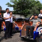 Man in a cap sits in a blue-and-orange pedicab while four uniformed men chat beside a row of orange rickshaws in a sunny parking lot.