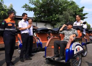 Man in a cap sits in a blue-and-orange pedicab while four uniformed men chat beside a row of orange rickshaws in a sunny parking lot.