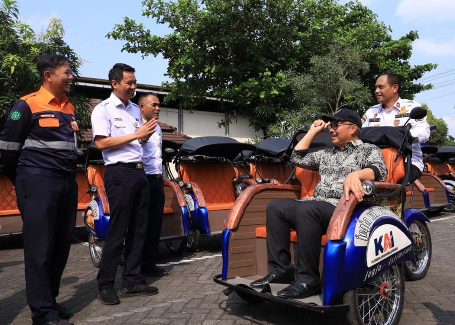 Man in a cap sits in a blue-and-orange pedicab while four uniformed men chat beside a row of orange rickshaws in a sunny parking lot.