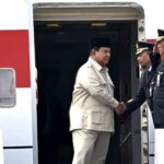 Two men shake hands at the open door of a red-and-white airplane, one in a beige suit and black cap, the other in a navy pilot uniform with gold badges.