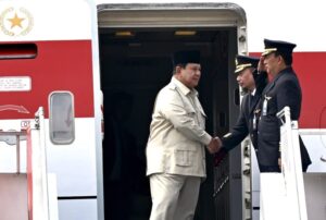 Two men shake hands at the open door of a red-and-white airplane, one in a beige suit and black cap, the other in a navy pilot uniform with gold badges.