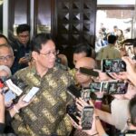 Man in a batik shirt speaks to reporters surrounded by microphones and smartphones in a crowded lobby corridor in front of a wooden door.