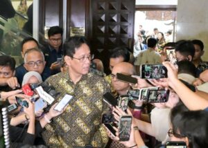Man in a batik shirt speaks to reporters surrounded by microphones and smartphones in a crowded lobby corridor in front of a wooden door.