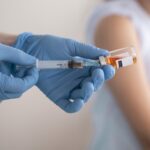 Medical worker in blue gloves draws vaccine from an amber vial into a syringe.