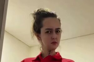 Close-up of a person with dark hair tied back, wearing a red shirt, looking to the side in an indoor setting.