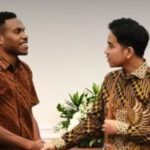Two men in batik shirts shake hands in a formal setting, with flowers in the background.