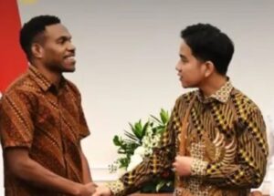 Two men in batik shirts shake hands in a formal setting, with flowers in the background.