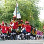 Parade of people in red uniforms marching with drums and a flag down a tree-lined street with spectators nearby.