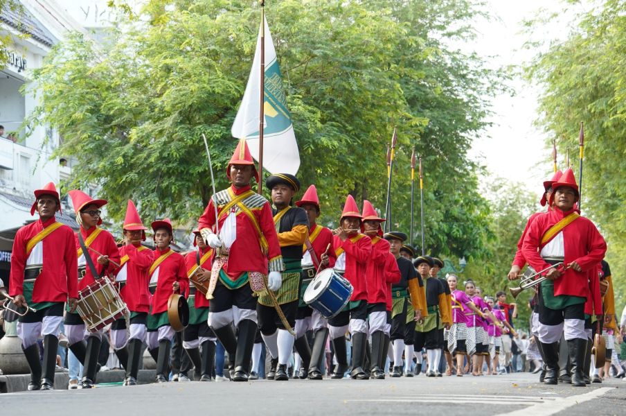 Parade of people in red uniforms marching with drums and a flag down a tree-lined street with spectators nearby.