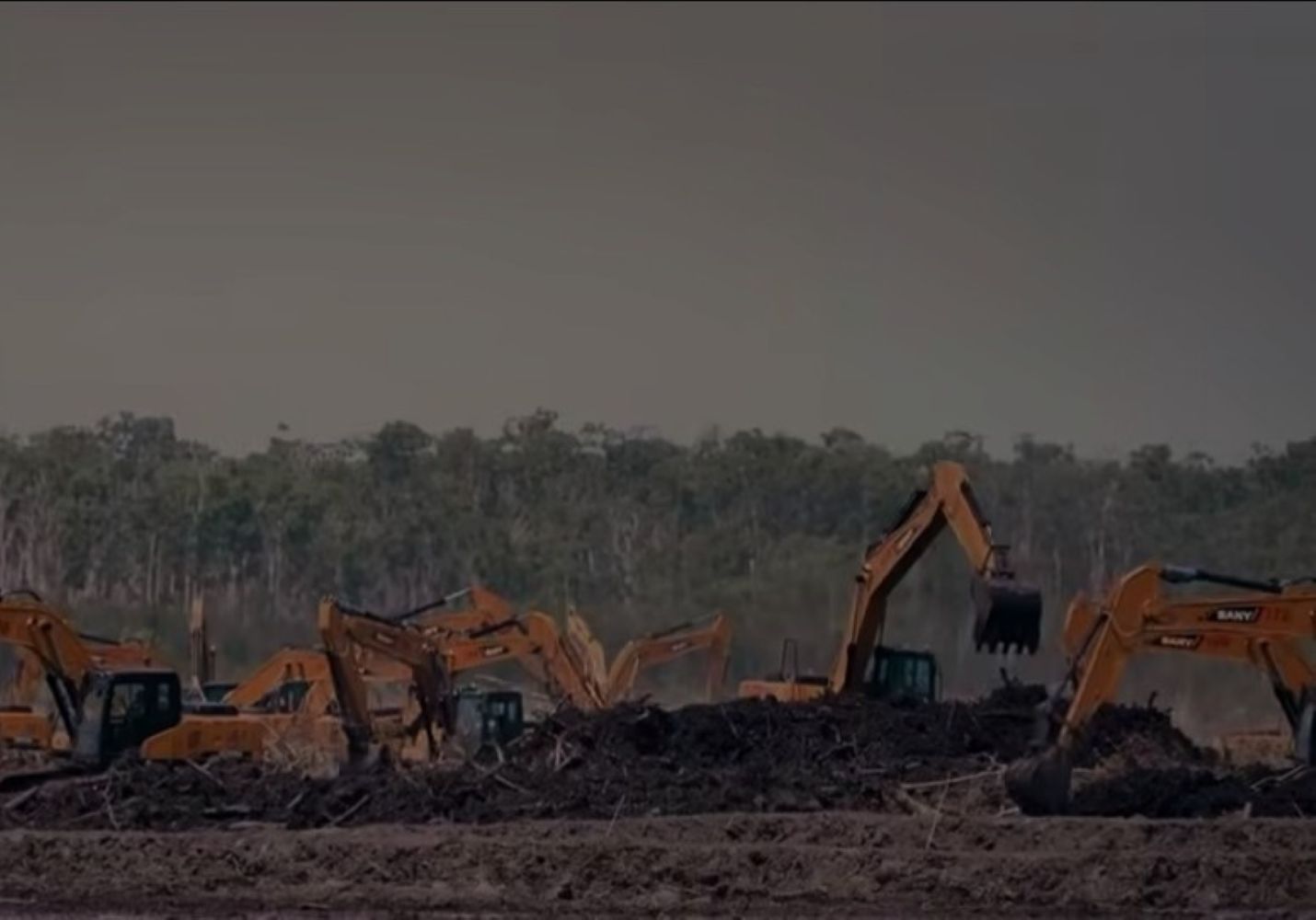 Several orange excavators work on a dirt site with a large pile of debris in the foreground, trees in the background.