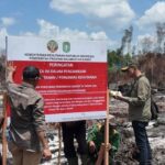 Officials in green uniforms inspecting a large red-and-white warning sign at a burned forest site, with caution tape and scorched ground in the background.