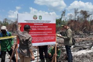 Officials in green uniforms inspecting a large red-and-white warning sign at a burned forest site, with caution tape and scorched ground in the background.