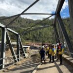 Group of construction workers in hard hats and yellow vests stand on a partially completed steel bridge, with a forested hillside in the background.