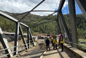 Group of construction workers in hard hats and yellow vests stand on a partially completed steel bridge, with a forested hillside in the background.