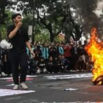 A young man in black speaks into a microphone as a pile of tires burns on a street protest, with a large crowd gathered behind him.