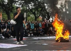 A young man in black speaks into a microphone as a pile of tires burns on a street protest, with a large crowd gathered behind him.
