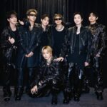 Seven men in black leather outfits posing together backstage against a dark curtain.