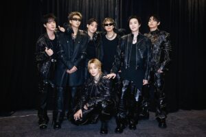 Seven men in black leather outfits posing together backstage against a dark curtain.