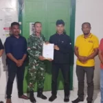 Six men pose indoors as a soldier in camouflage hands a document to a young man in black; others watch the handover.