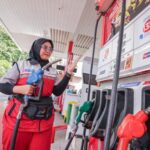 Female gas station attendant in a red uniform and hijab holding a fuel nozzle while gesturing at a pump in a bright station setting.