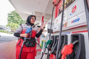 Female gas station attendant in a red uniform and hijab holding a fuel nozzle while gesturing at a pump in a bright station setting.