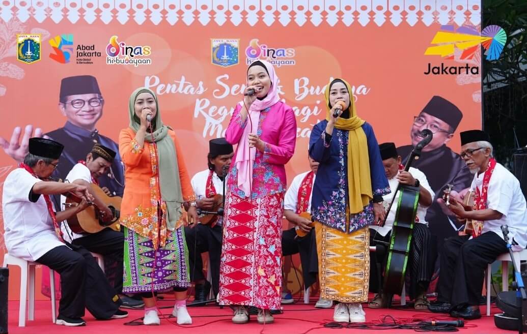 Group of four women in colorful traditional outfits singing on a stage with musicians behind them at a cultural event banner in Jakarta