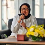 Woman wearing a black hijab and glasses speaks into a microphone during a panel, with yellow flowers on the table nearby.