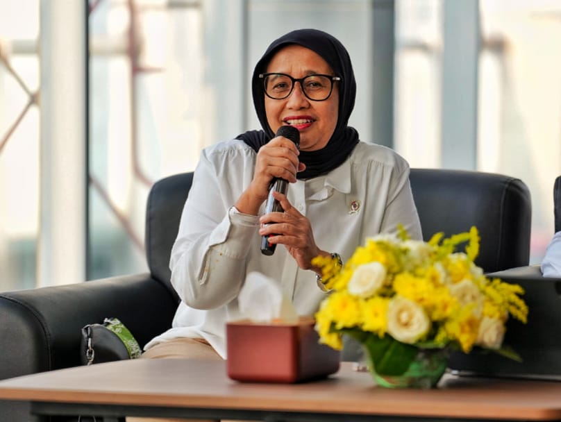 Woman wearing a black hijab and glasses speaks into a microphone during a panel, with yellow flowers on the table nearby.