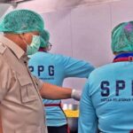 People in hairnets and face masks work at a food-preparation station, a supervisor in a tan uniform observes two staff wearing blue SPPG shirts beside a metal prep area.