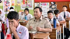 Man in a khaki uniform seated at a desk in a classroom, surrounded by students in white shirts and ties.