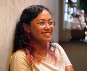 Smiling woman with glasses sits against a textured wall in a warm indoor setting.