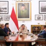 Group of officials sit around a polished wooden conference table in a formal meeting, with the Indonesian flag behind them and framed portraits on the wall behind the center speaker.