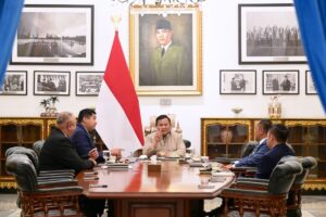 Group of officials sit around a polished wooden conference table in a formal meeting, with the Indonesian flag behind them and framed portraits on the wall behind the center speaker.