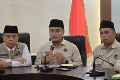 Three men in beige uniforms and black peci hats sit at a conference table during a formal briefing, with an Indonesian flag in the background and microphones in front of them.